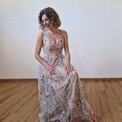 Frau in langem, floral besticktem Kleid sitzt auf Holzboden vor weißer Wand.