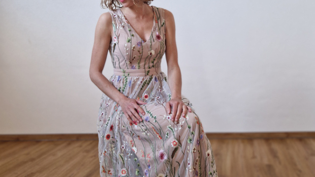 Frau in langem, floral besticktem Kleid sitzt auf Holzboden vor weißer Wand.