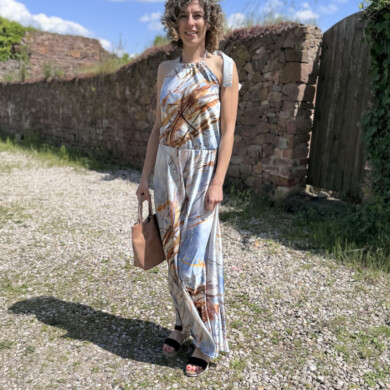 Frau in Sommerkleid mit Tasche vor alter Steinmauer bei Sonnenschein.