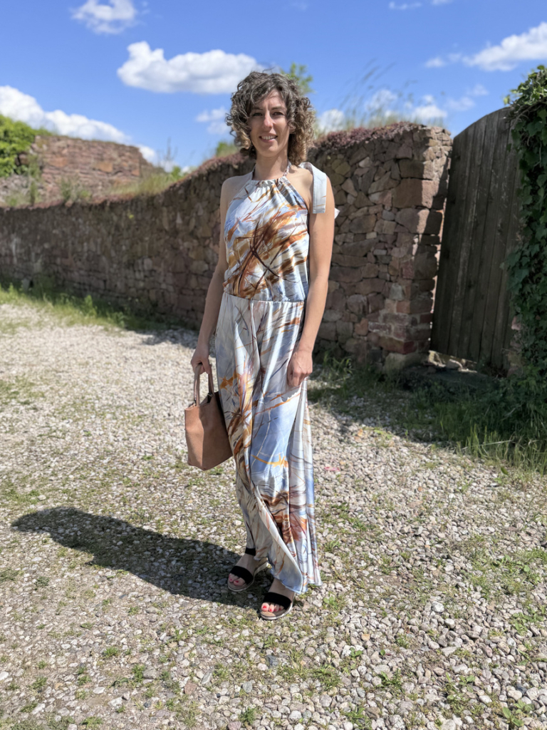 Frau in Sommerkleid mit Tasche vor alter Steinmauer bei Sonnenschein.